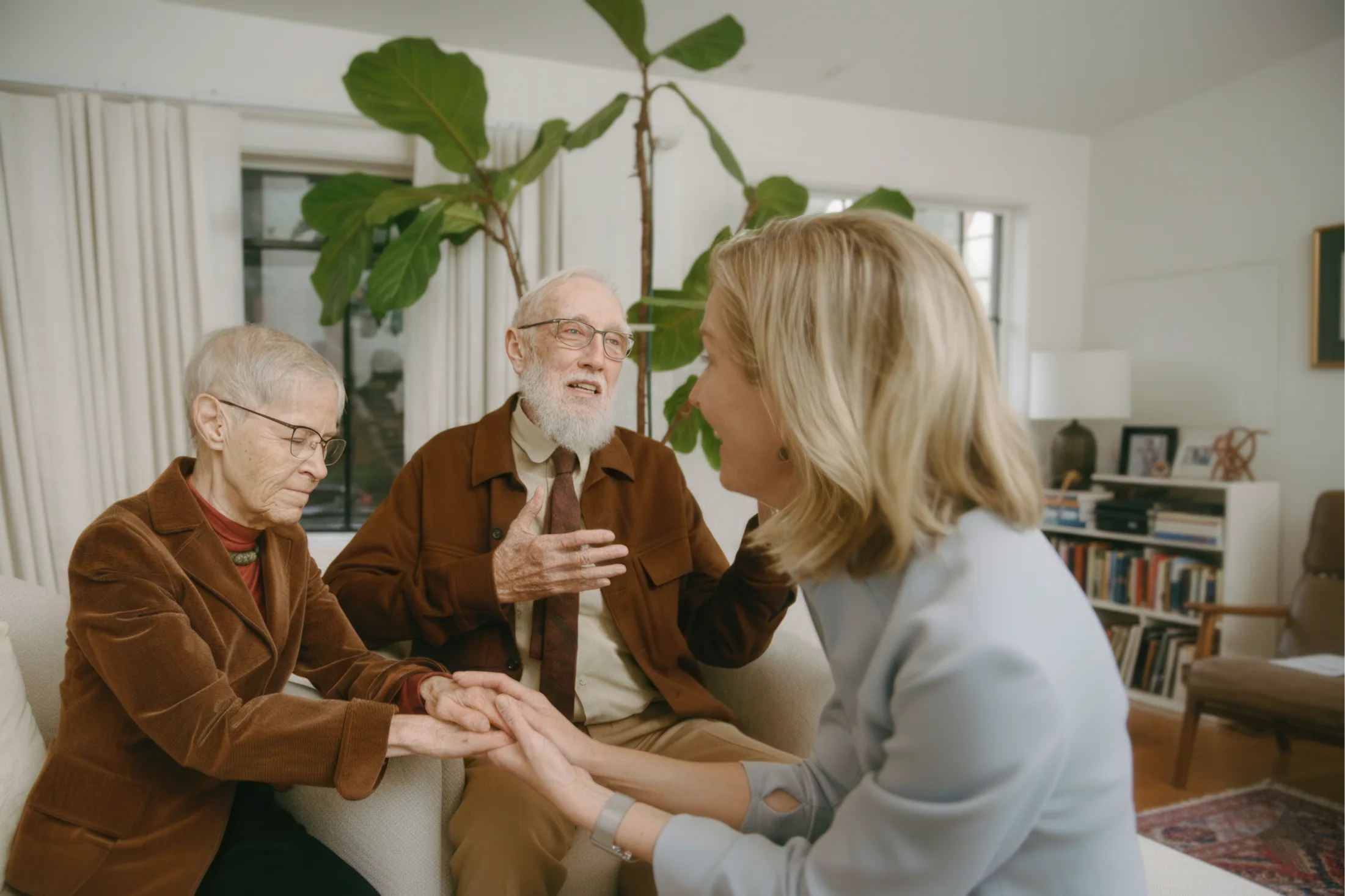 Caregiver holding hands with senior couple in bright living room during supportive conversation.