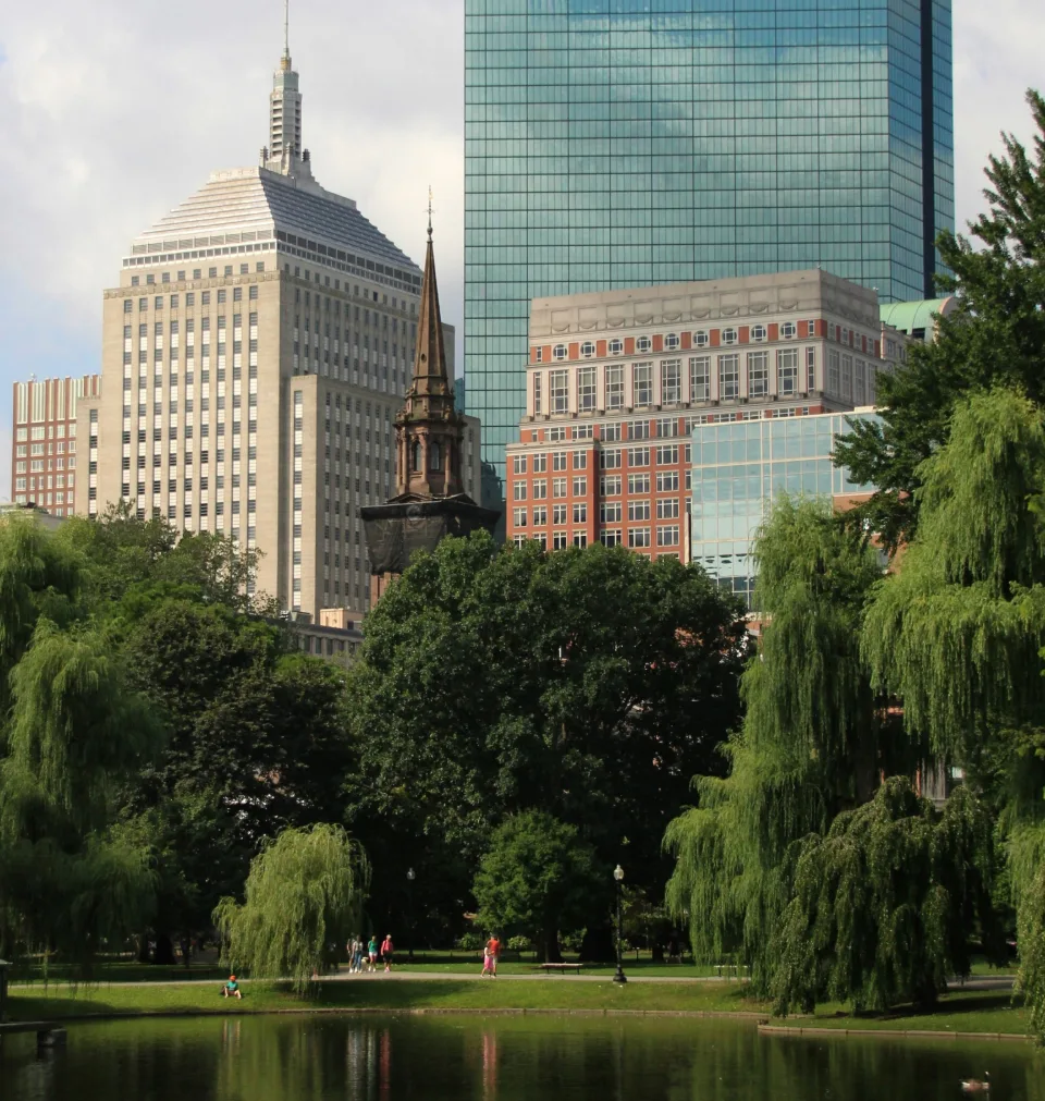 Urban park with pond, willow trees, and Boston skyline featuring historic and modern buildings.