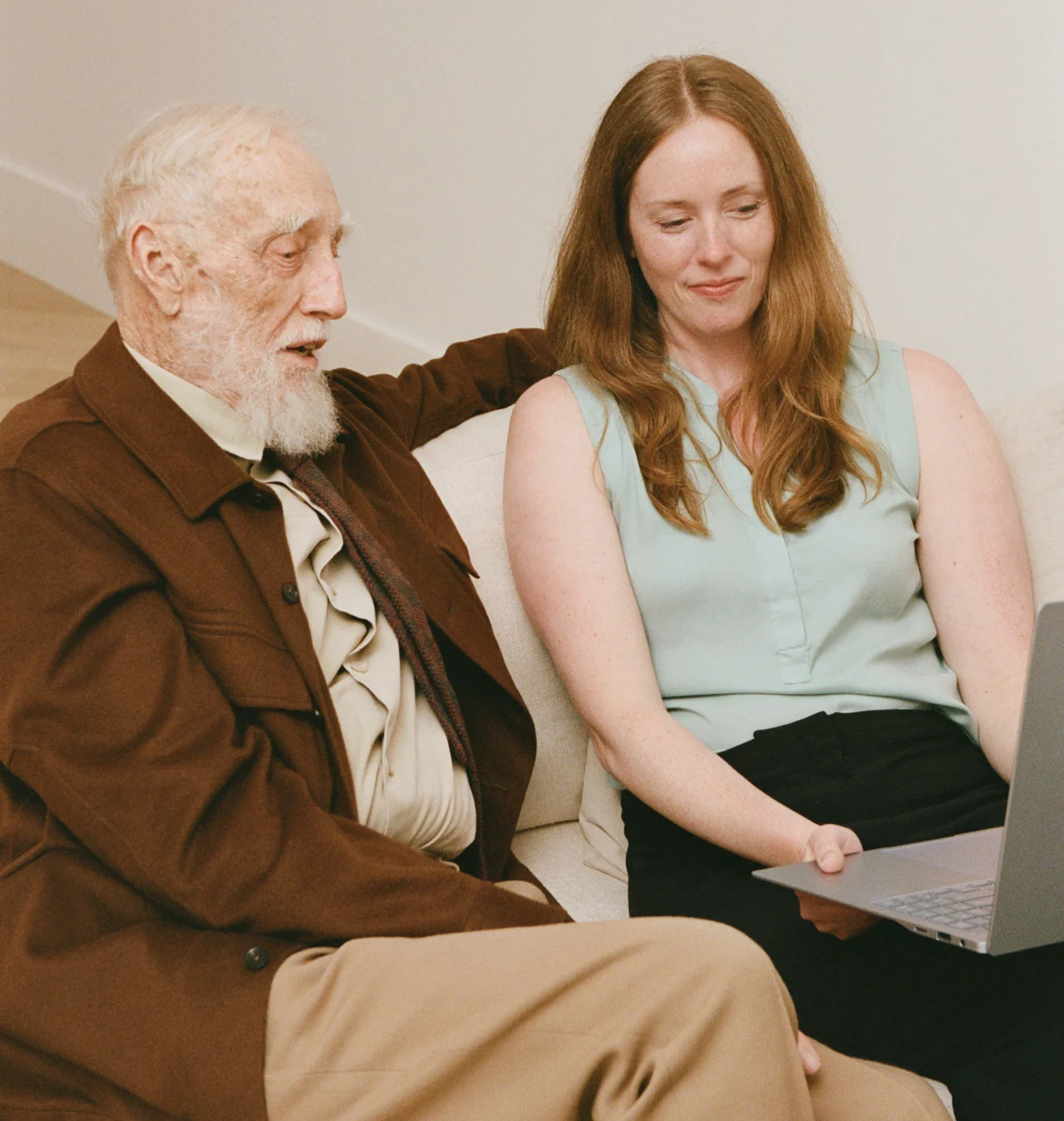 Senior man in brown jacket with caregiver reviewing laptop together, representing virtual care management services.