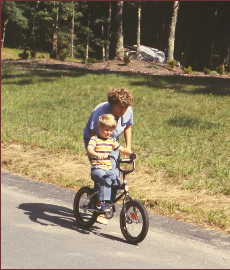 Parent helping a child learn to ride a bike on a neighborhood road