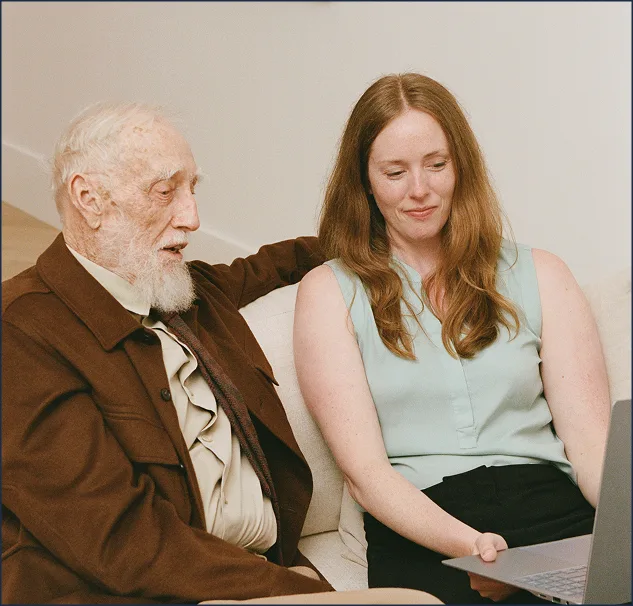 Older man in brown jacket with caregiver reviewing information on laptop together.