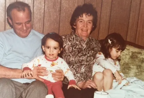 Older man and woman sitting with two young children on a bed, smiling at camera in a warm family moment.