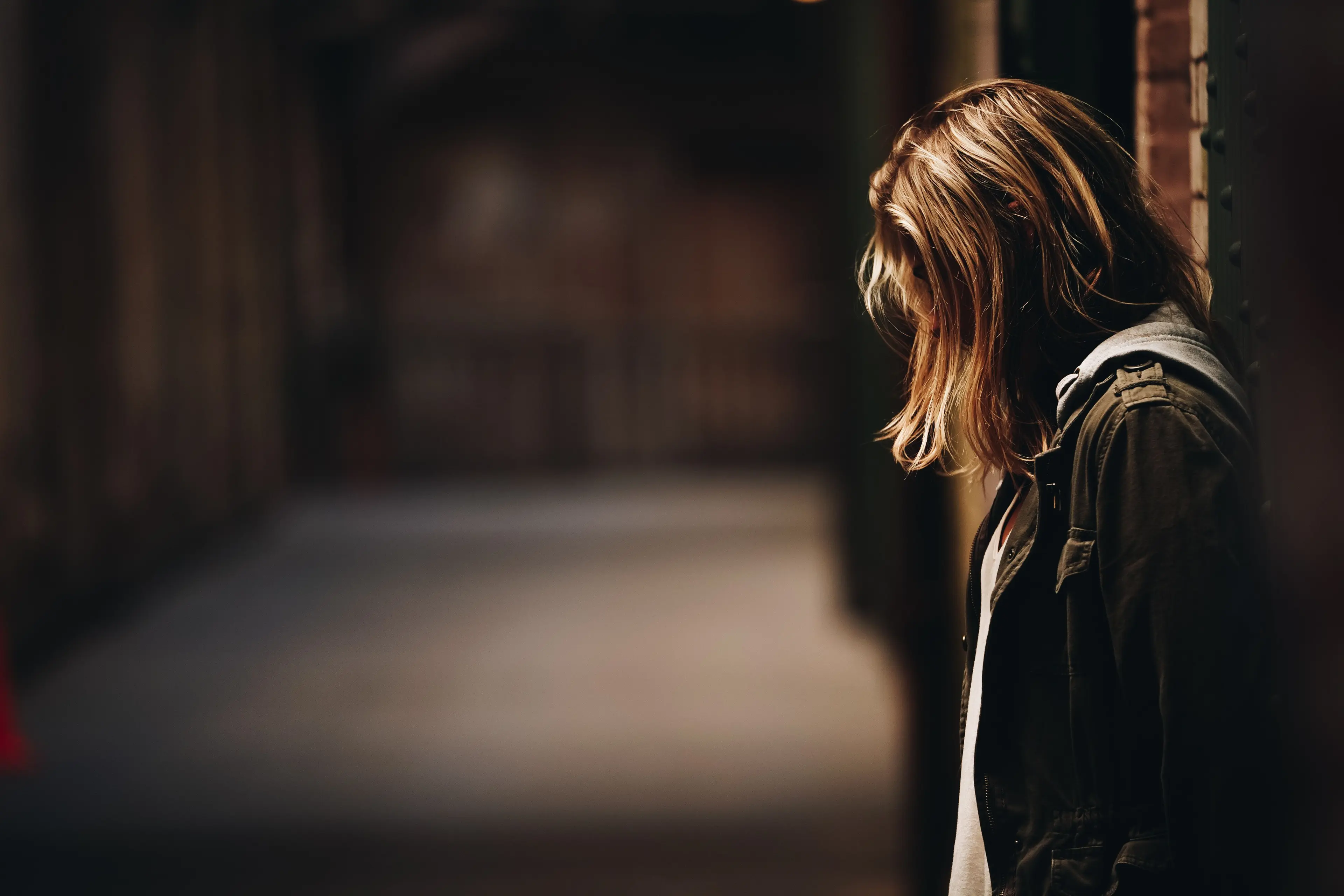 A woman stands alone in a dimly lit hallway, leaning against a wall with her head bowed, appearing sad or deep in thought.