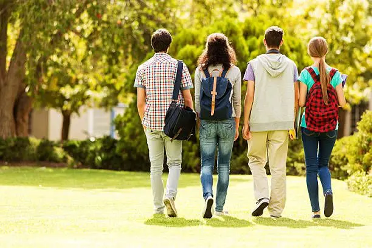 Four teenagers walk together outdoors on a sunny day, carrying backpacks and talking as they head across a grassy area.