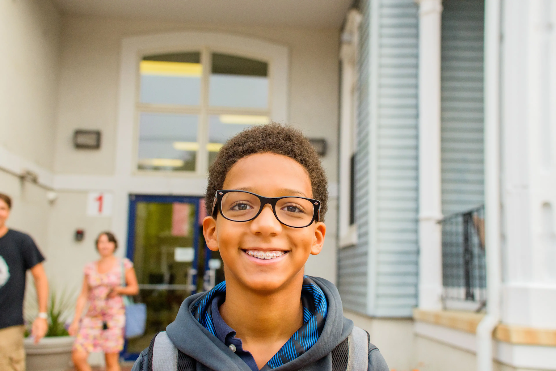 A smiling student wearing glasses and a backpack stands outside a school building, with other people in the background.