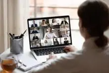 A person sits at a desk using a laptop for a video conference, with multiple people visible on the screen.
