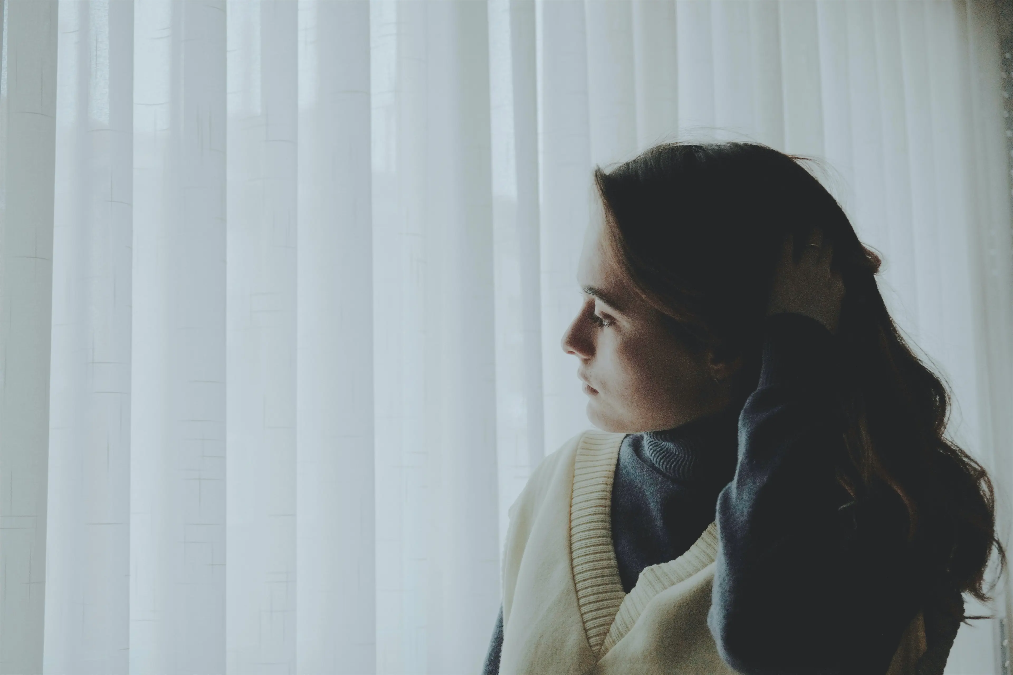 A woman stands indoors looking out a window with a thoughtful expression, one hand resting on her hair.