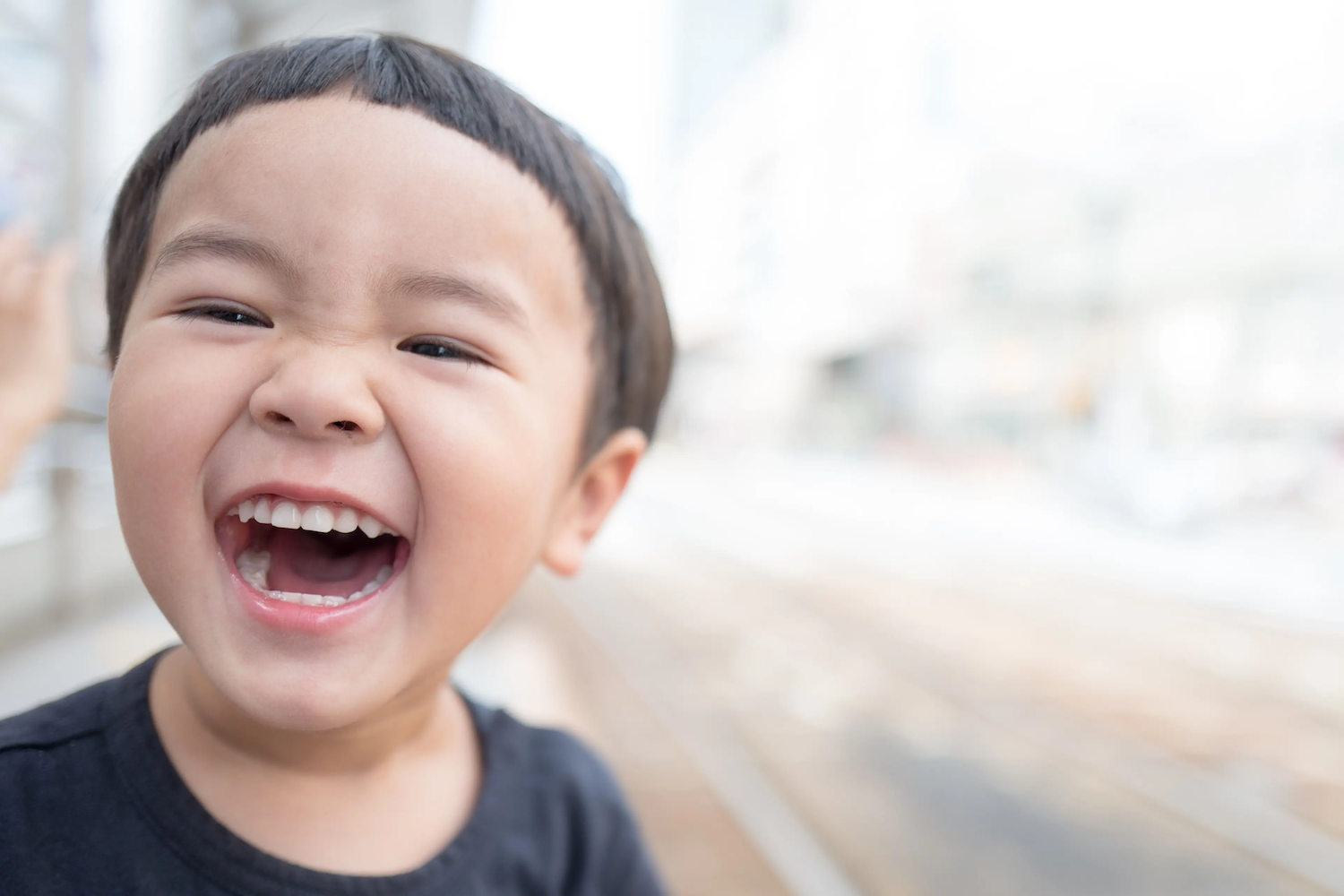 A young boy smiles widely and laughs outdoors, showing his teeth in a joyful expression.