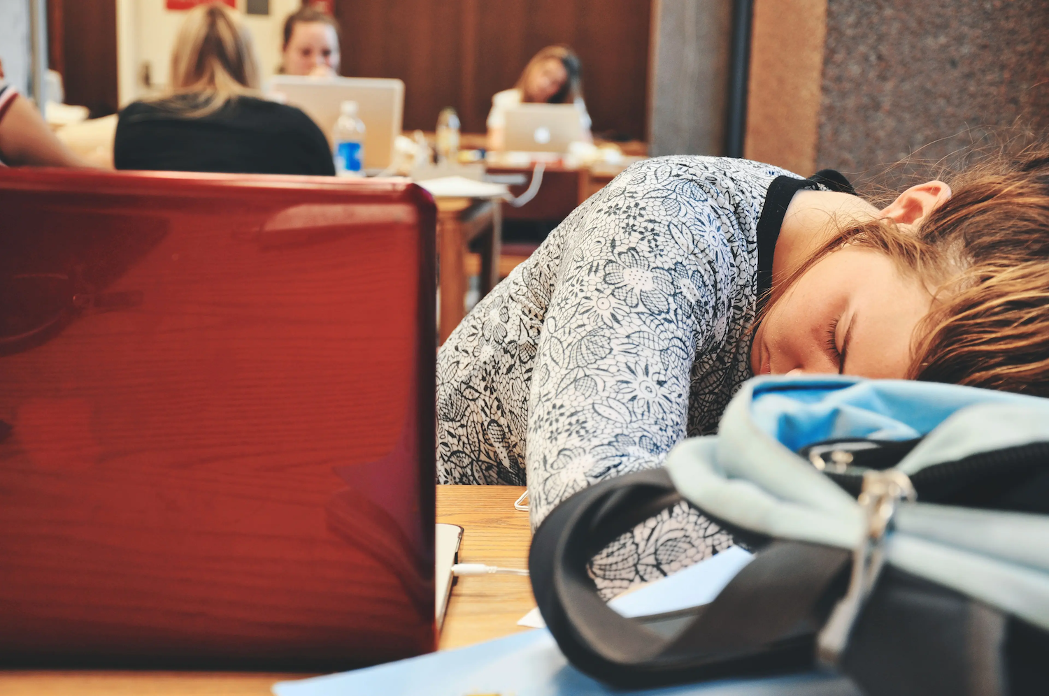A student rests their head on a desk beside a laptop and backpack in a classroom or study area, appearing tired.