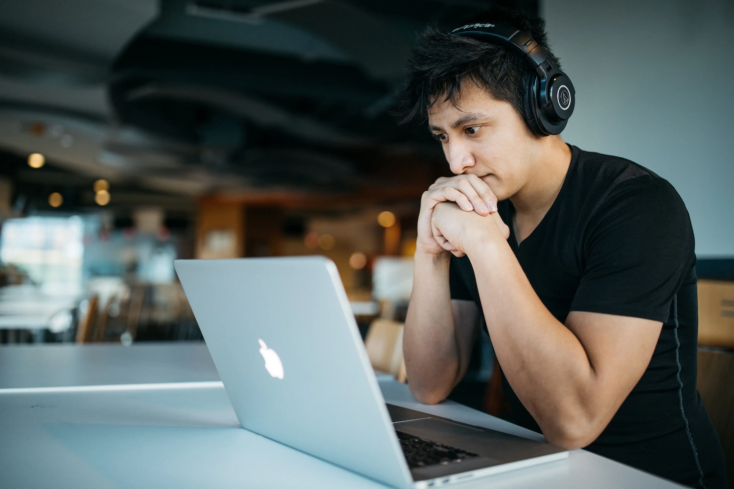 A man wearing headphones sits at a desk, looking intently at a laptop screen with his hands clasped under his chin.