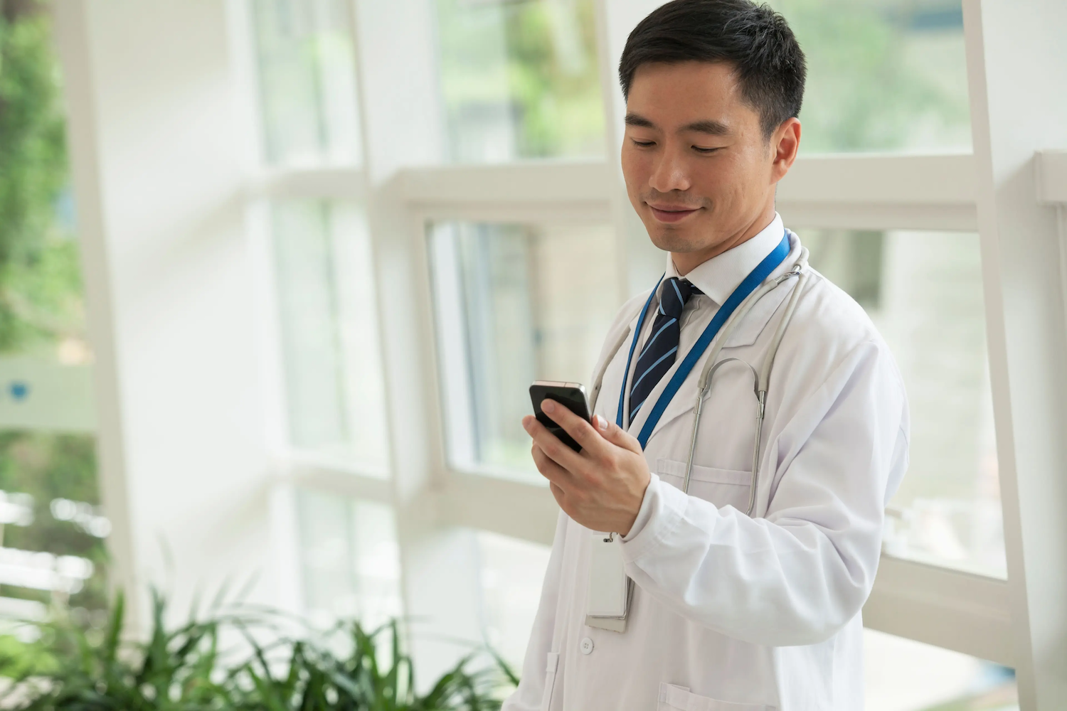 A doctor wearing a white coat and stethoscope looks at a smartphone with a slight smile while standing in a bright hallway.