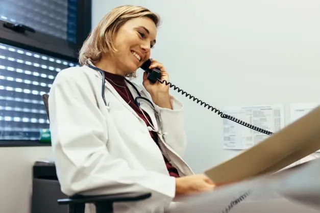 A smiling doctor wearing a white coat and stethoscope talks on the phone while holding a patient file in an office.