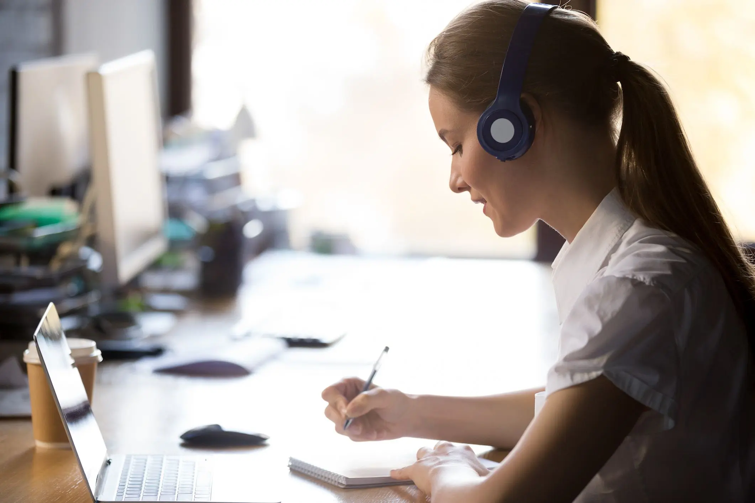 A woman wearing headphones sits at a desk, writing in a notebook while looking at a laptop screen.