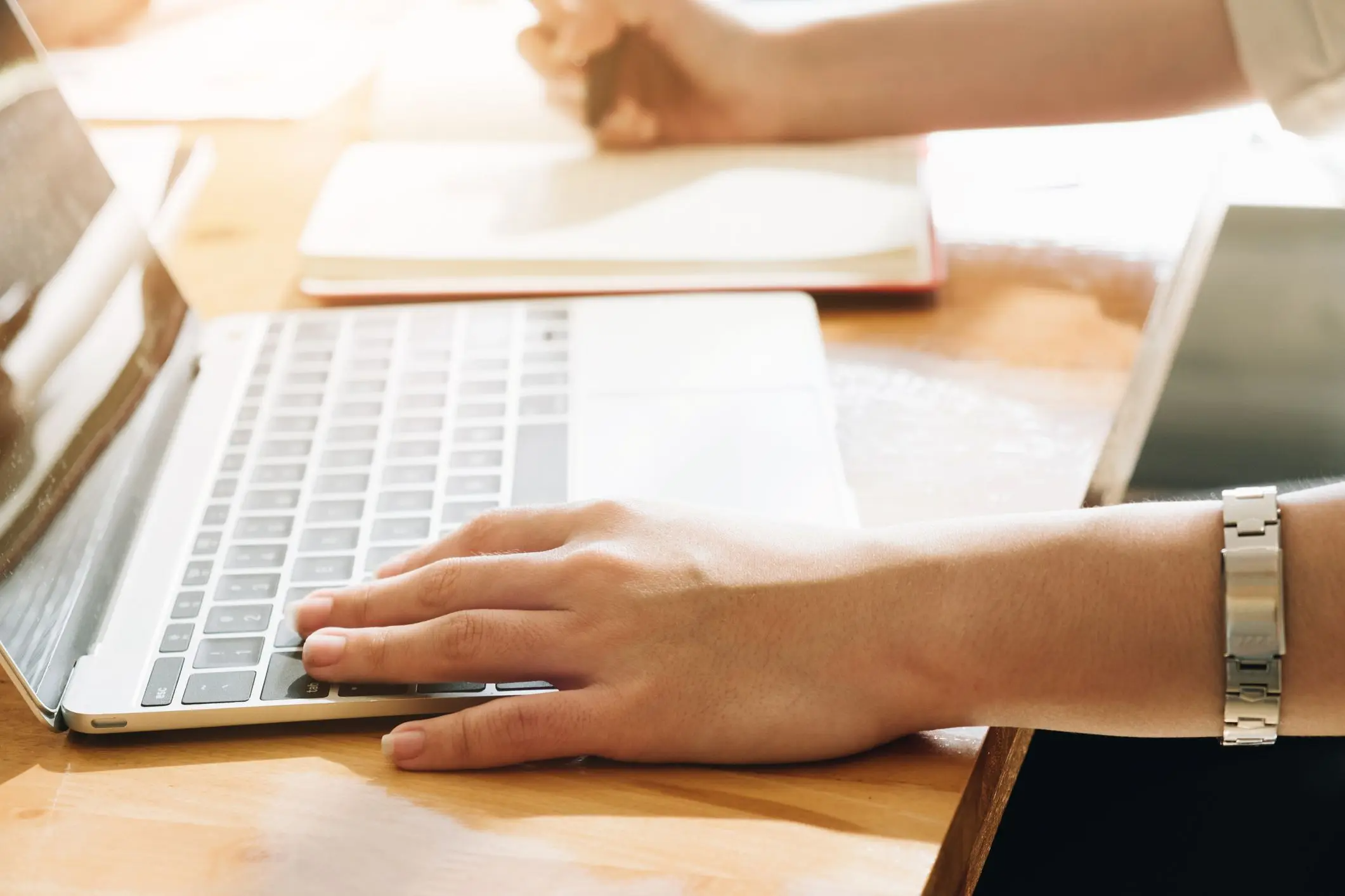 A person typing on a laptop on a wooden desk in bright light.