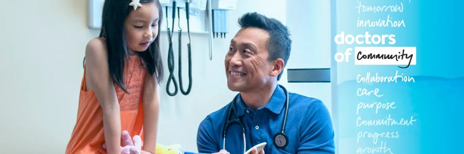 A smiling doctor in a blue shirt with a stethoscope talks with a young girl in an orange dress sitting on an exam table, with medical equipment in the background.
