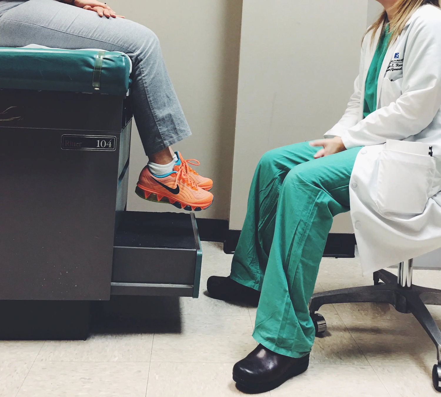A doctor in green scrubs and a white coat talks with a patient sitting on an exam table wearing gray pants and orange sneakers.