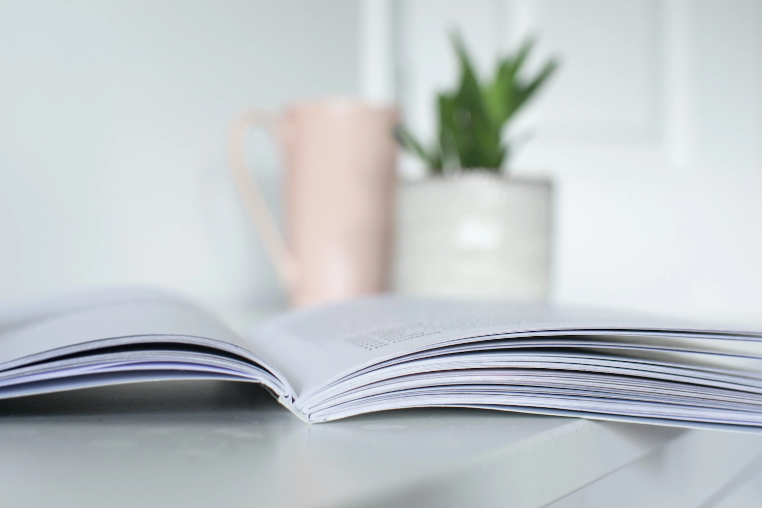 An open book lies on a white surface with a blurred pink mug and small potted plant in the background.