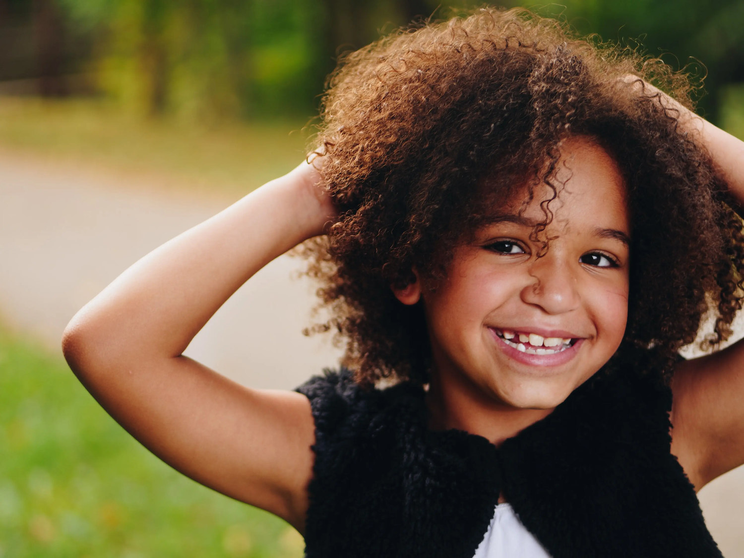 A smiling young child with curly hair holds their hands behind their head while standing outdoors on a sunny day.