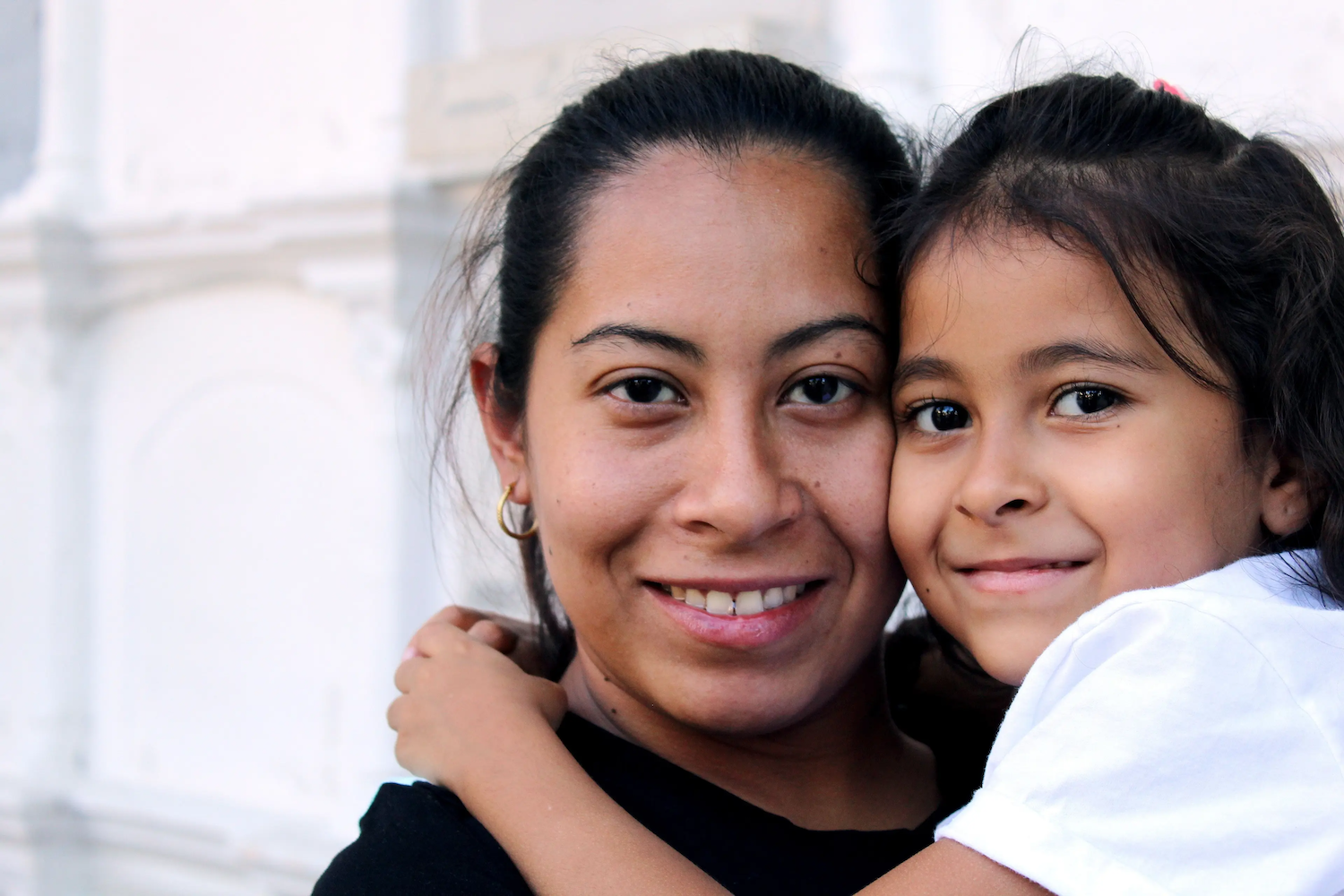 A smiling woman and a young girl share a close hug, both looking at the camera with warmth and affection.
