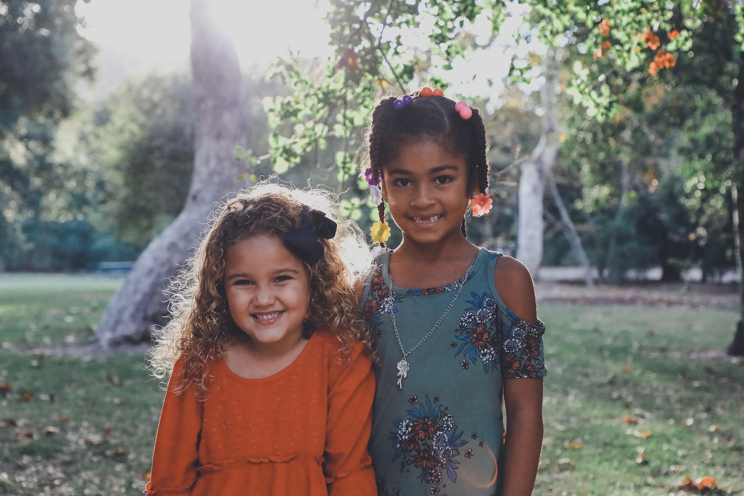Two young girls smiling outdoors in a sunlit park, one wearing an orange dress and the other a blue floral dress, standing close together in front of trees.