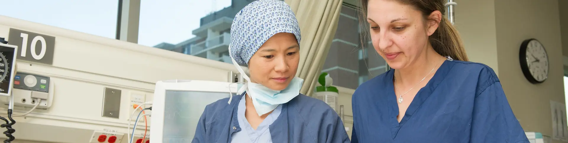 Two healthcare professionals in blue scrubs review information together in a hospital room, with medical equipment visible in the background.