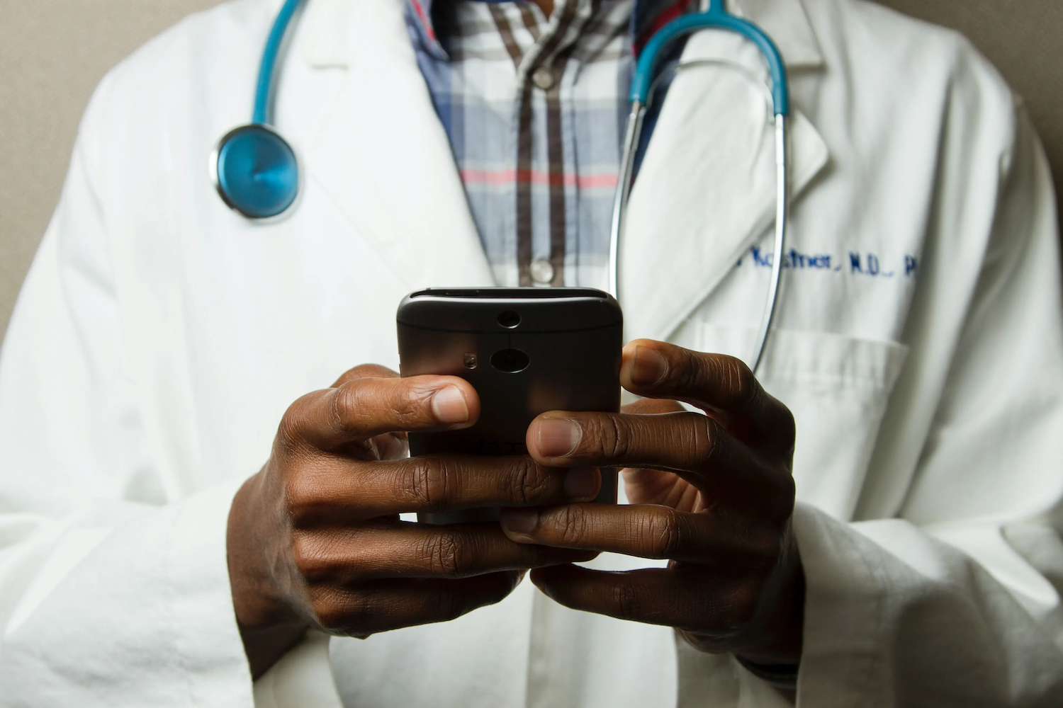 A doctor wearing a white coat and stethoscope uses a smartphone, focusing on their hands and the device.