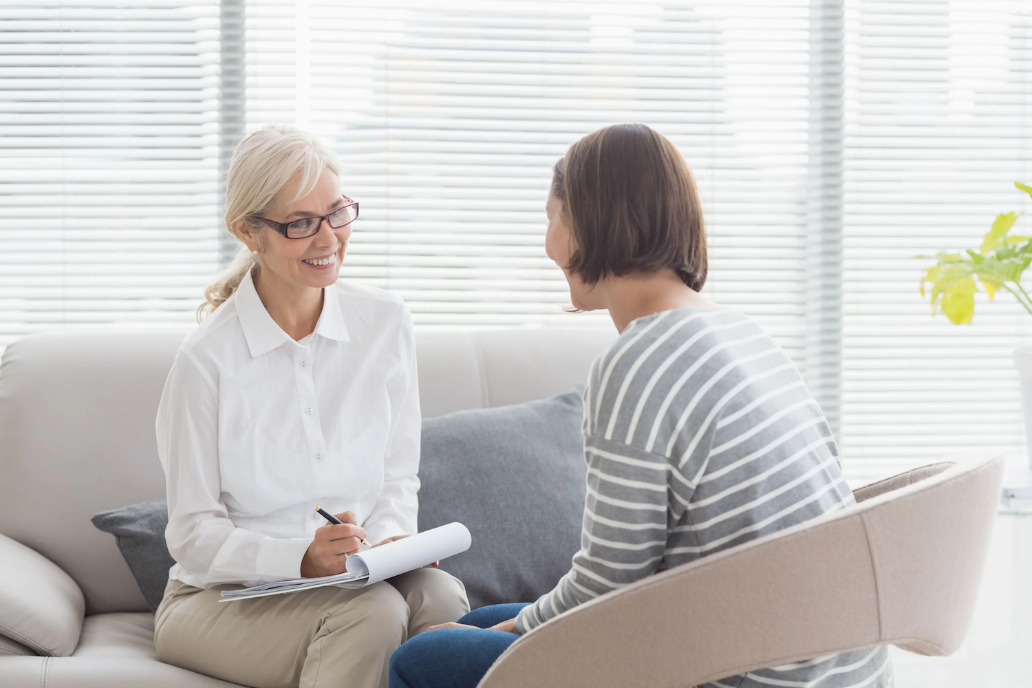 Smiling therapist taking notes while talking with a client in a bright, comfortable office setting.