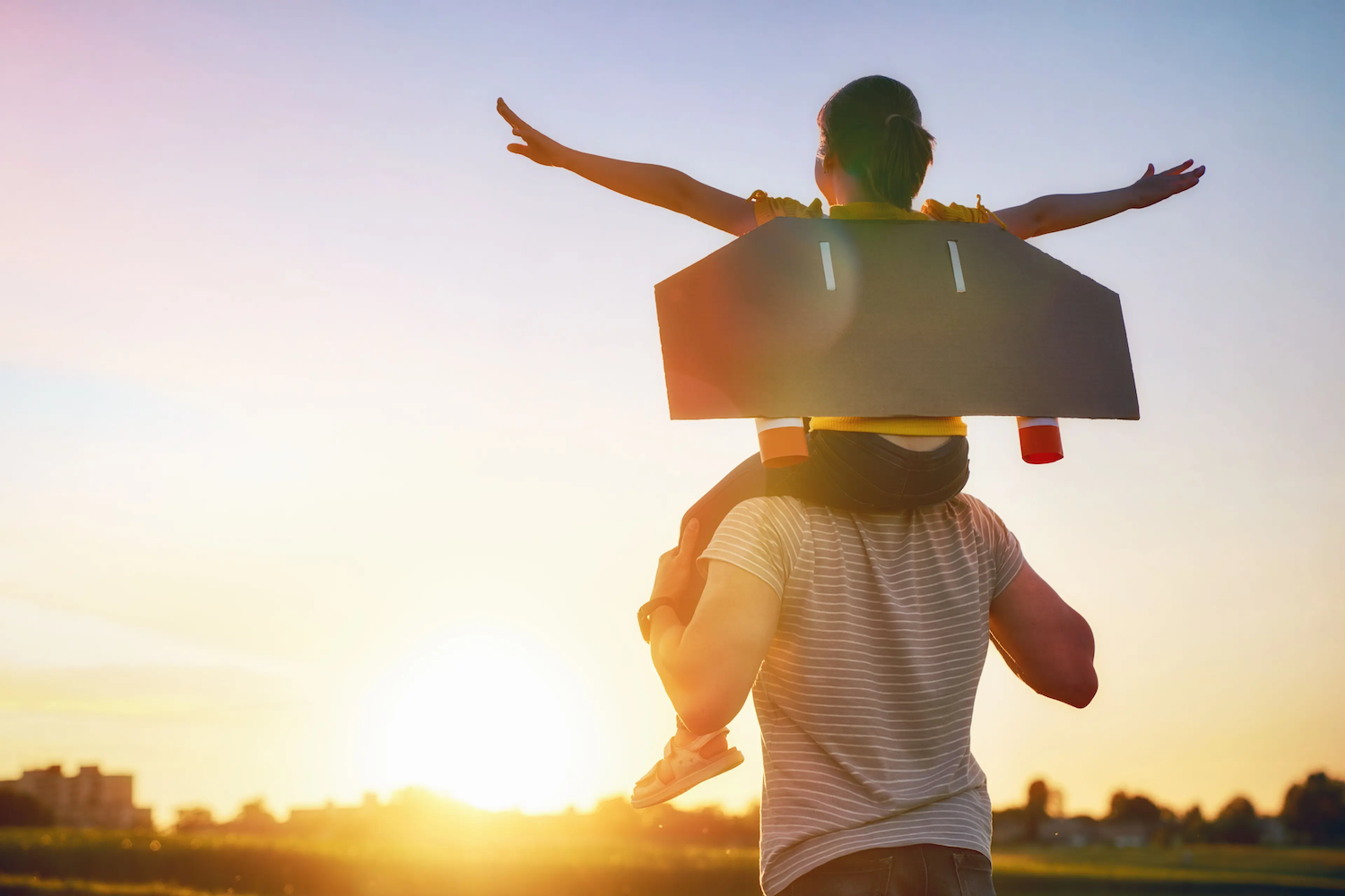 Child wearing a cardboard jetpack sitting on an adult’s shoulders with arms outstretched, enjoying the sunset in an open field.