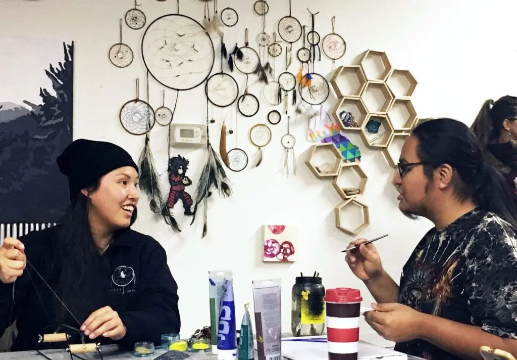 Two people sitting at a table engaged in a craft activity, surrounded by dreamcatchers and hexagonal wall shelves filled with small items.