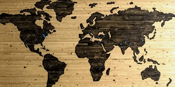 Wooden world map wall art with dark continents on a light wood background, decorated with small blue dots marking various locations.