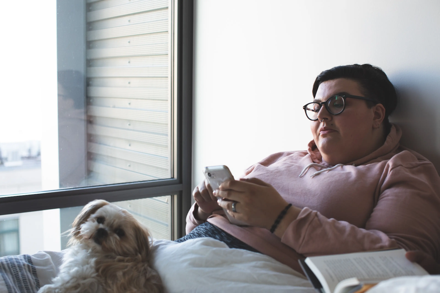 Person wearing glasses and a pink hoodie lying in bed near a window, holding a smartphone, with a small fluffy dog resting beside them and an open book nearby.