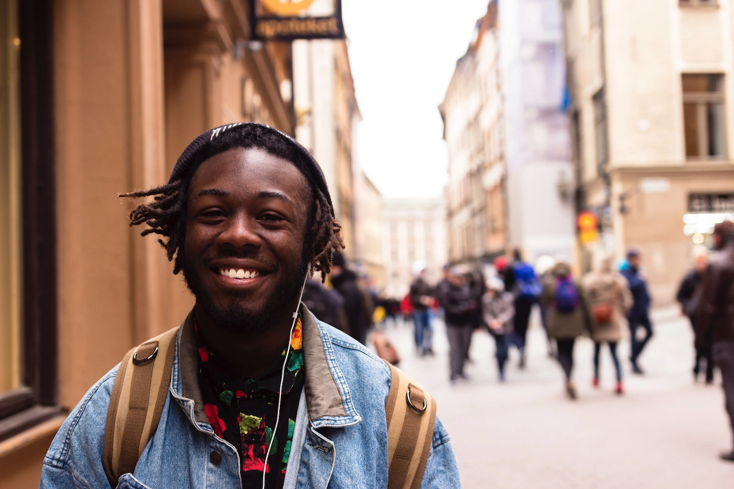 Smiling person wearing a denim jacket, colorful shirt, and backpack standing on a busy city street with blurred pedestrians in the background.