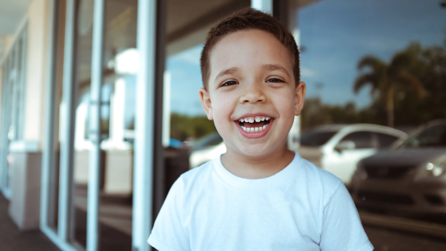 Smiling young boy wearing a white shirt standing outside near a row of windows, with cars and trees visible in the background.