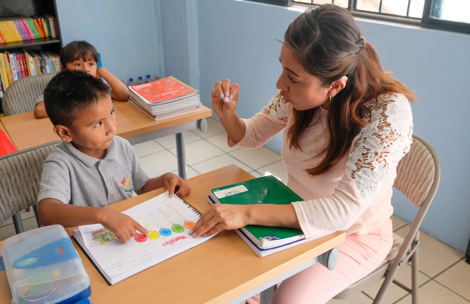Teacher sitting at a desk helping a young student with a colorful workbook in a classroom, while another child sits at a nearby desk in the background.