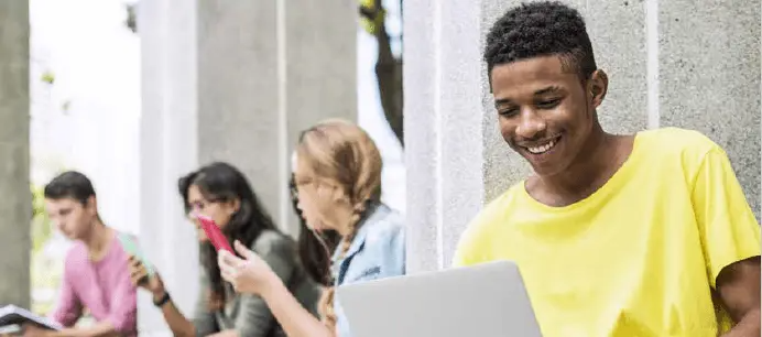 Group of college students sitting outdoors, with one smiling student in a yellow shirt using a laptop while others use their phones.