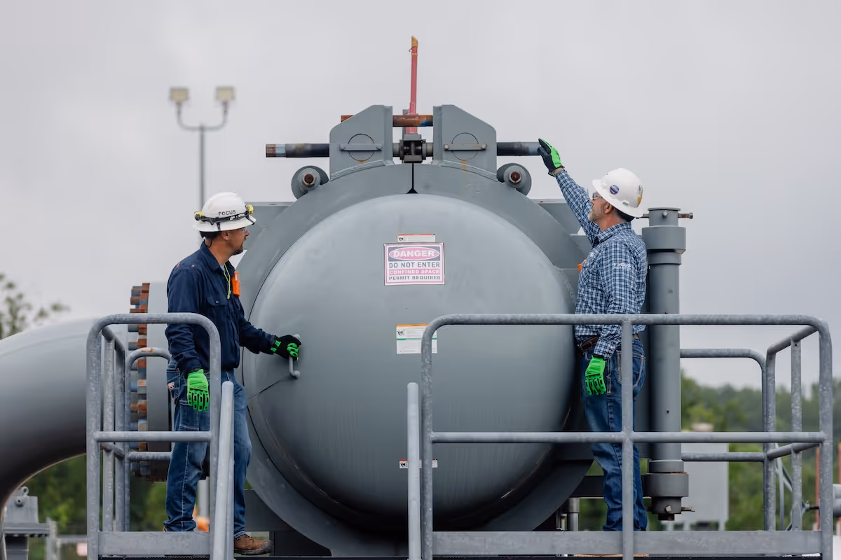 Two workers in hard hats and safety gloves standing on a platform next to a large pipeline vessel