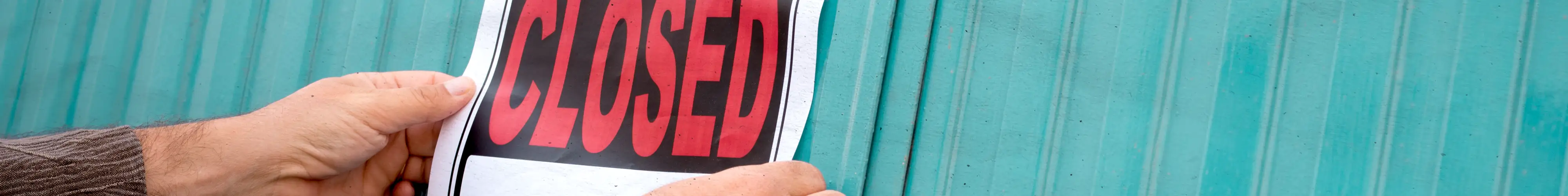 Hands hanging a red "Closed" sign on a teal wall.