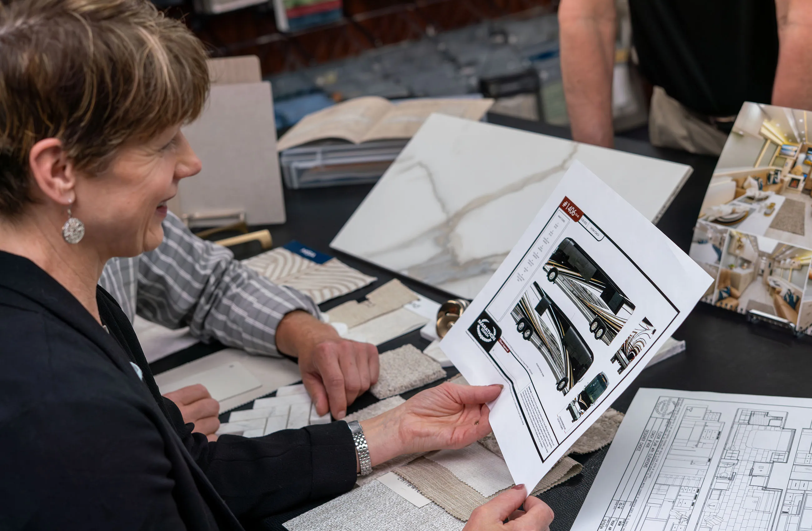 Two people reviewing design plans and fabric samples for a luxury RV with interior photos and floor plans on a table.