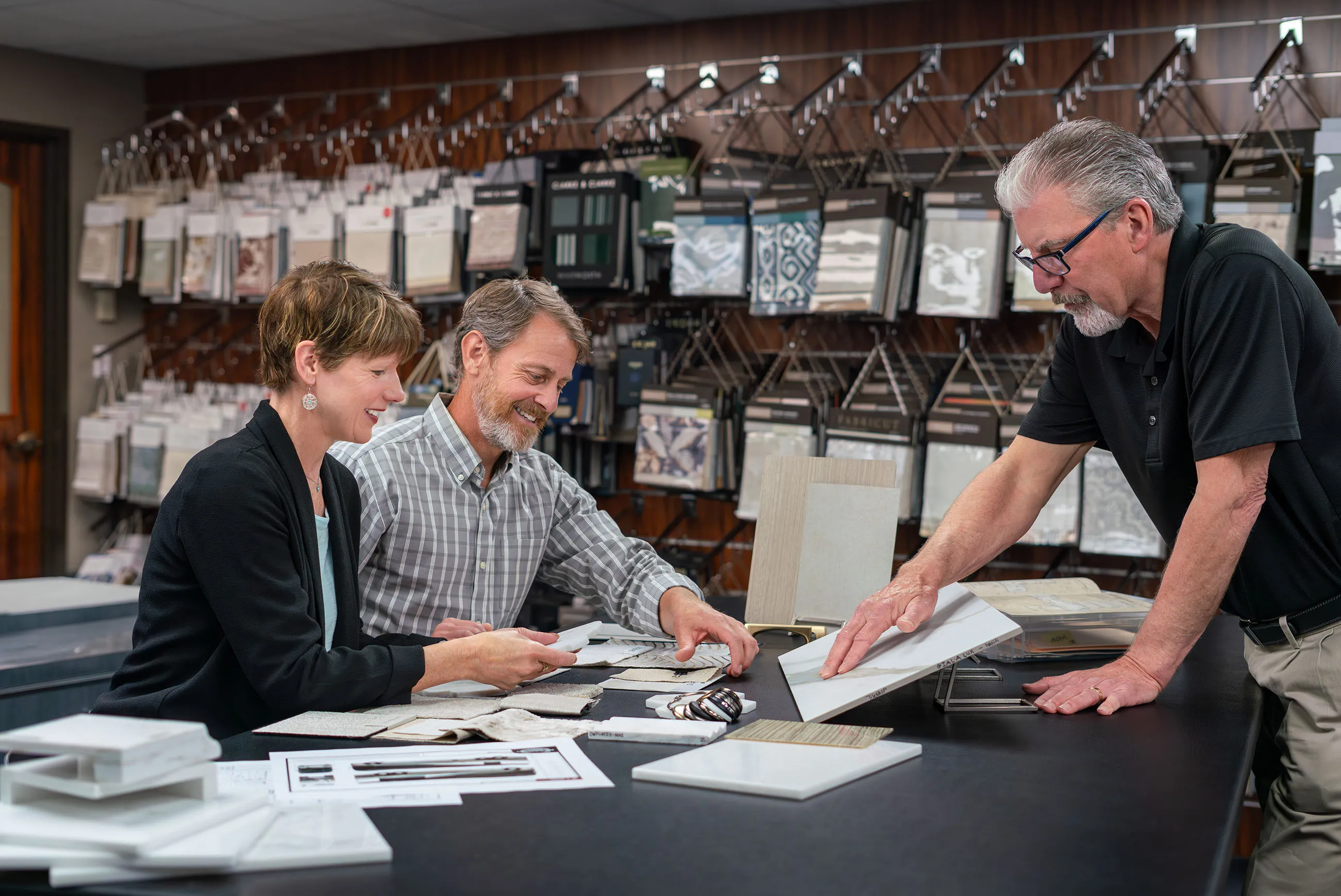 Two customers and a salesperson reviewing fabric and tile samples in a showroom with fabric swatches hanging on the wall.