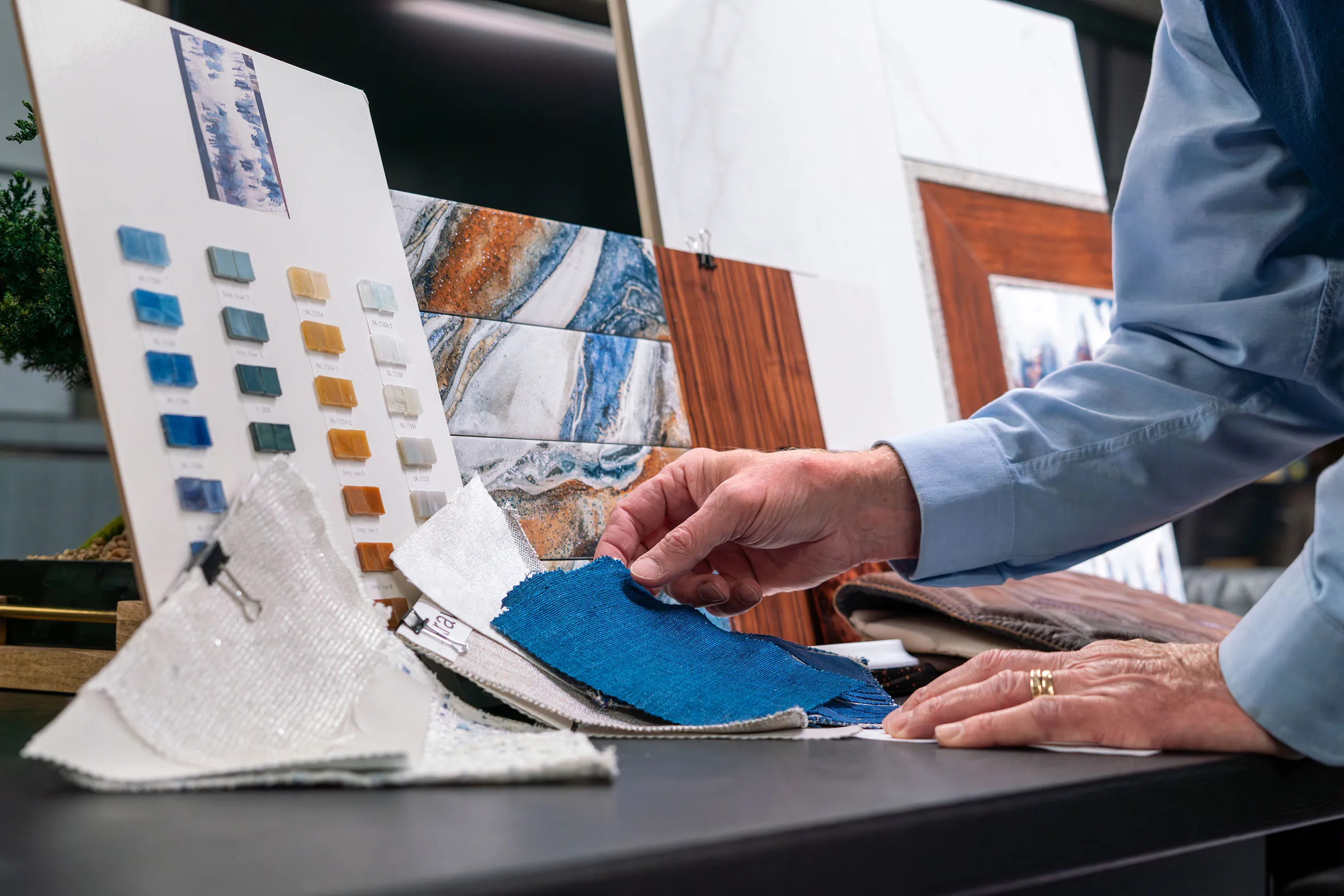 Person examining fabric swatches and color samples on a table with tile and wood samples in the background.