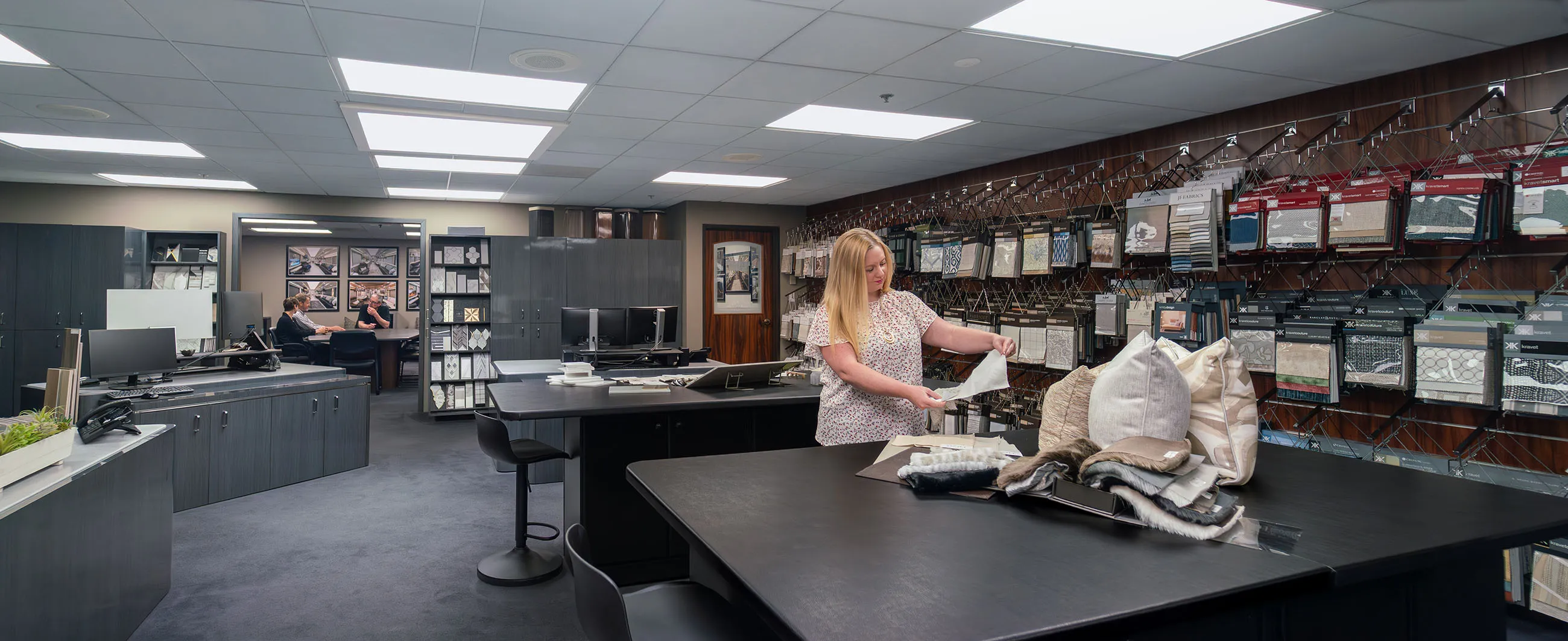 Interior of a fabric and design showroom with a woman examining fabric samples at a table filled with pillows and swatches, and three people meeting in a conference room in the background.