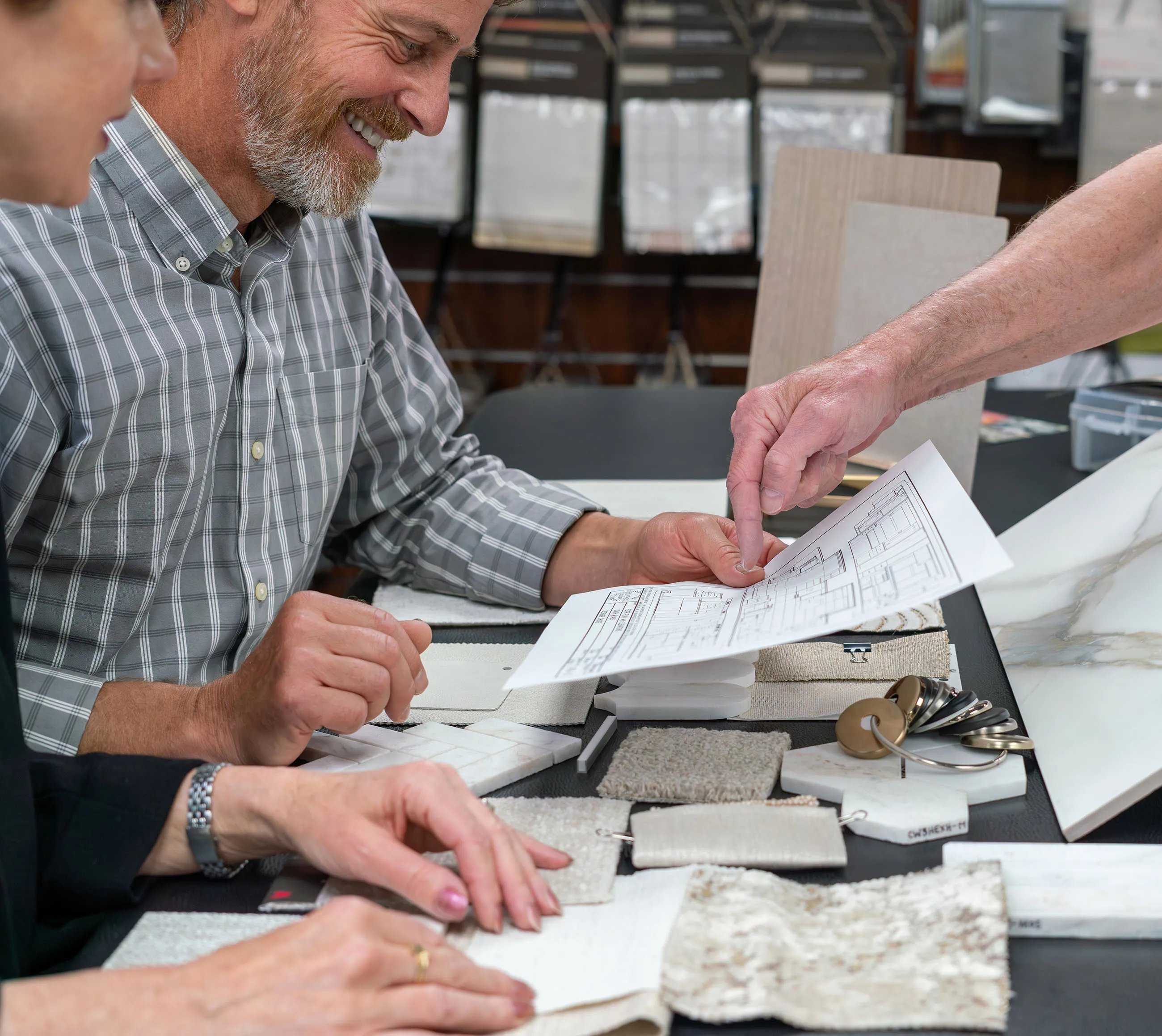 Three people reviewing fabric swatches, material samples, and a floor plan blueprint on a table.