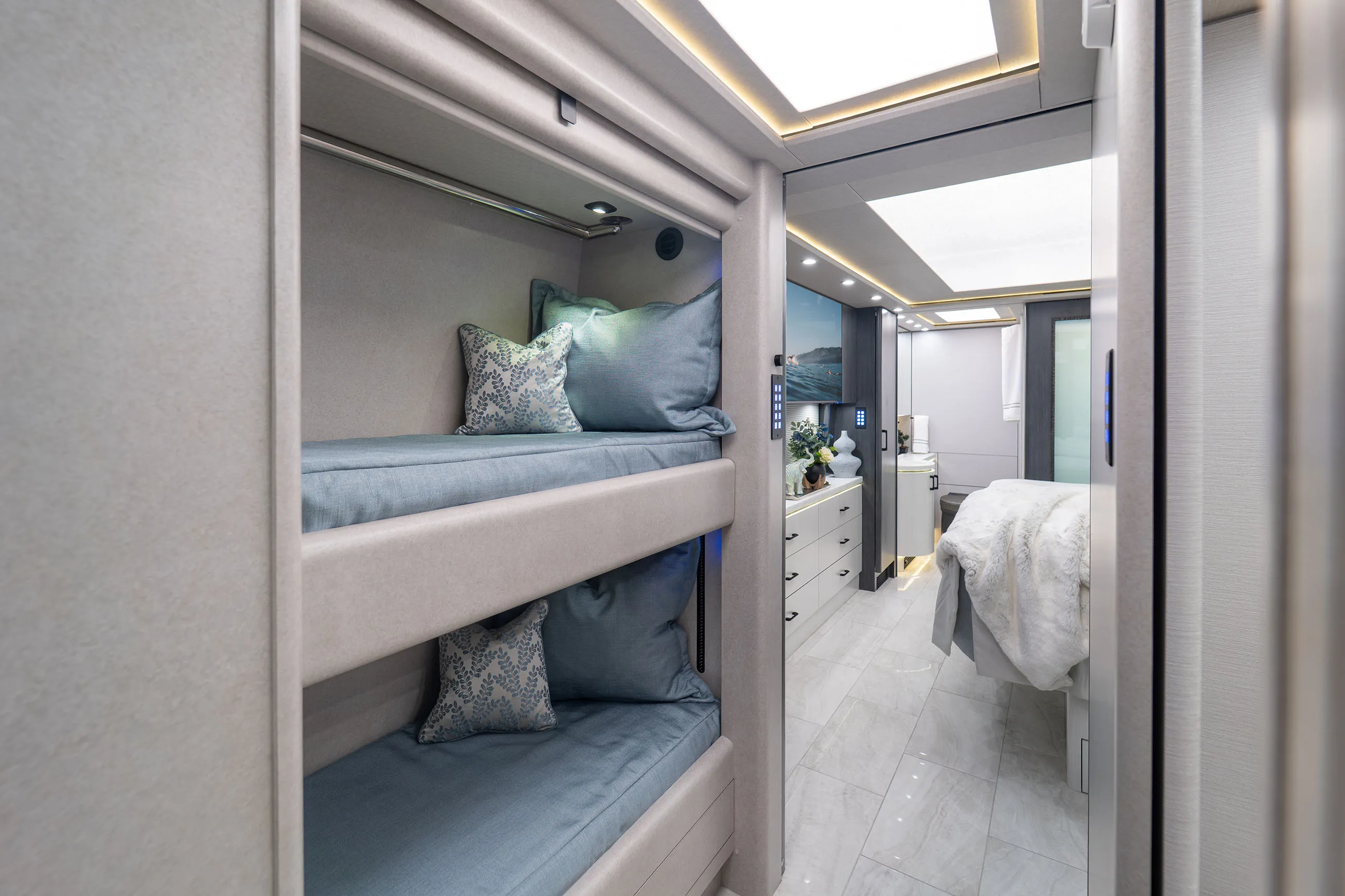 Modern RV interior featuring bunk beds with blue bedding and decorative pillows, hallway with drawers, and a bedroom with a white bedspread.