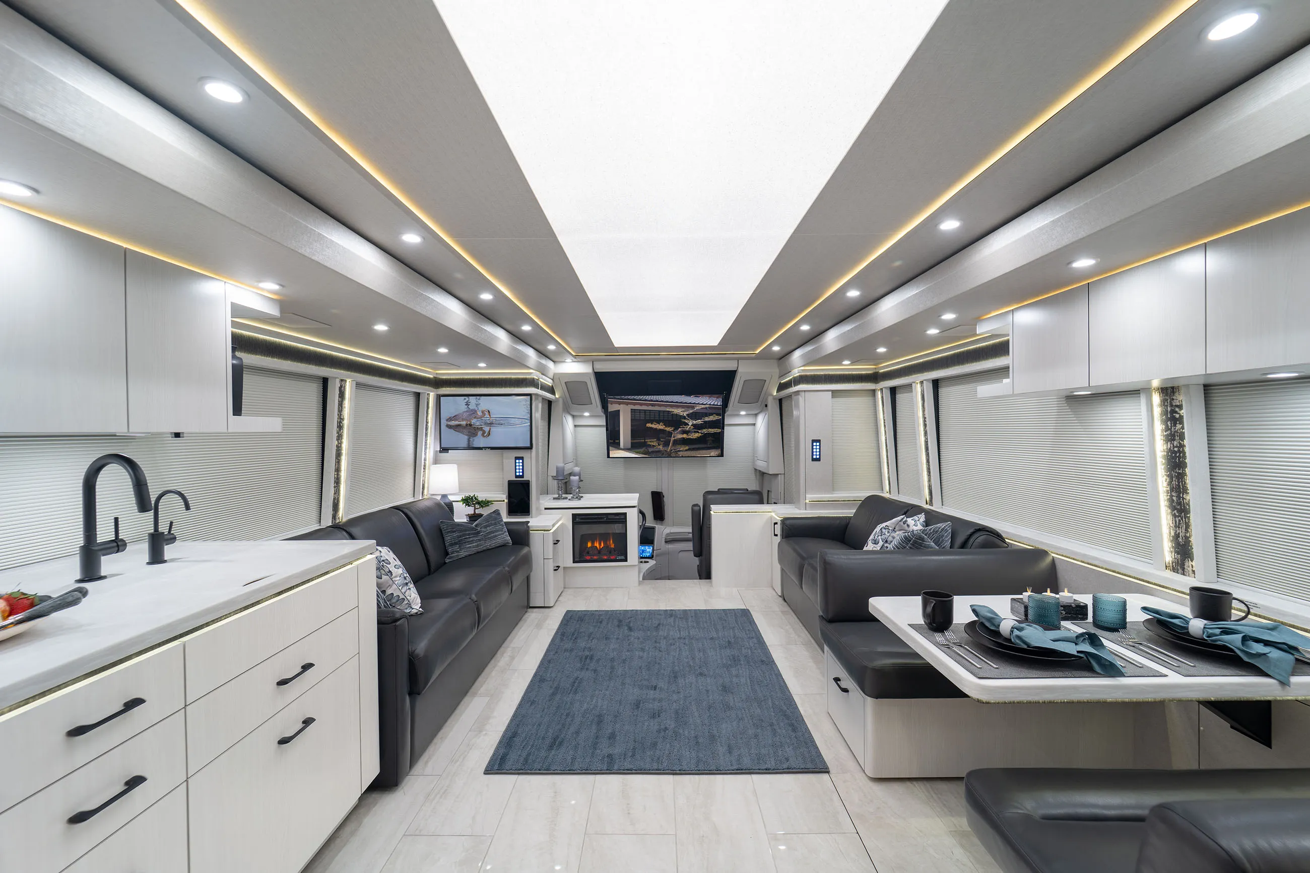 Modern RV interior with two black leather sofas, a dining area with table settings, kitchen with white cabinets, and a flat-screen TV above an electric fireplace.