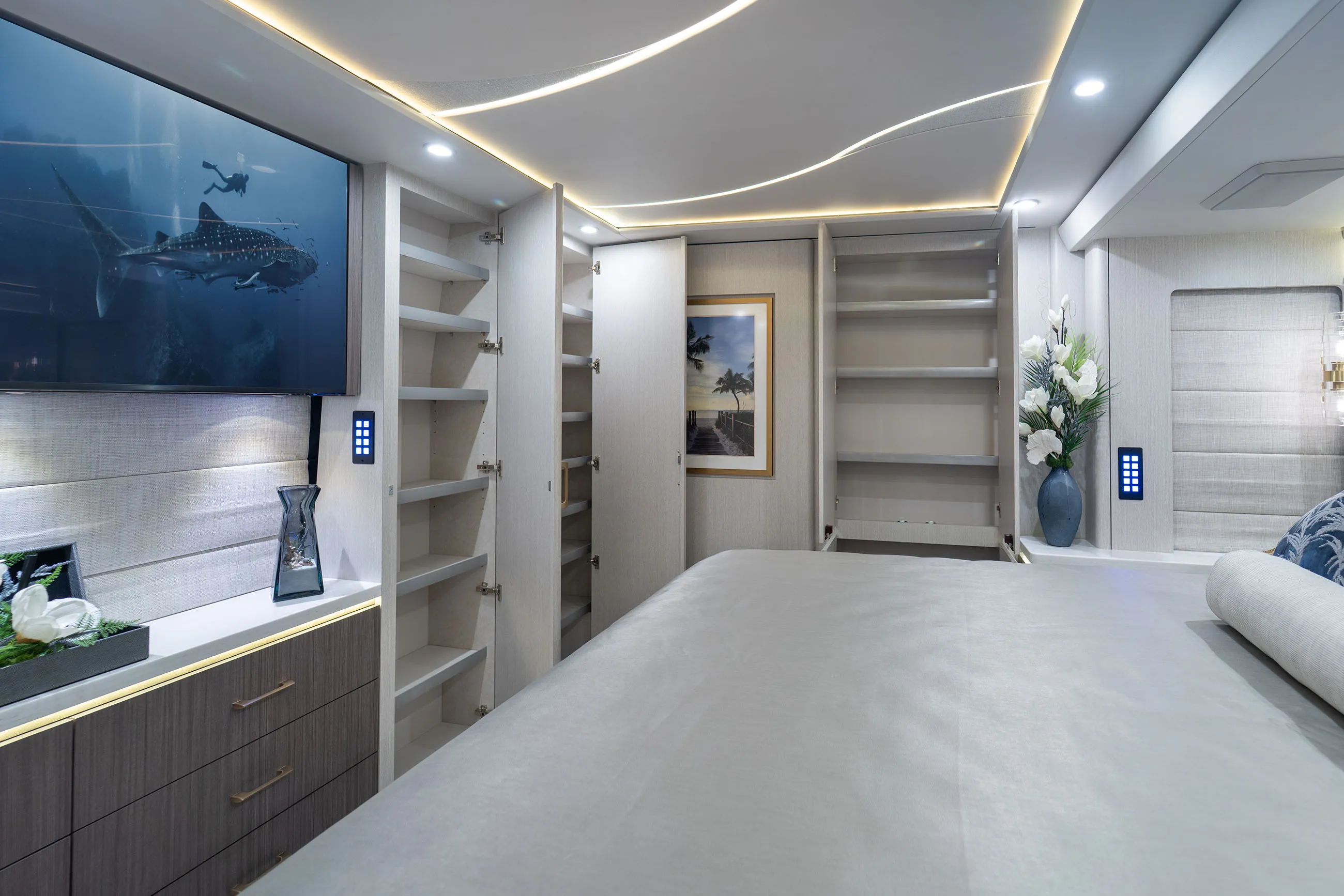 Modern bedroom interior with a large bed, open closets, wall-mounted TV showing underwater scene, decorative flowers, and framed beach artwork.