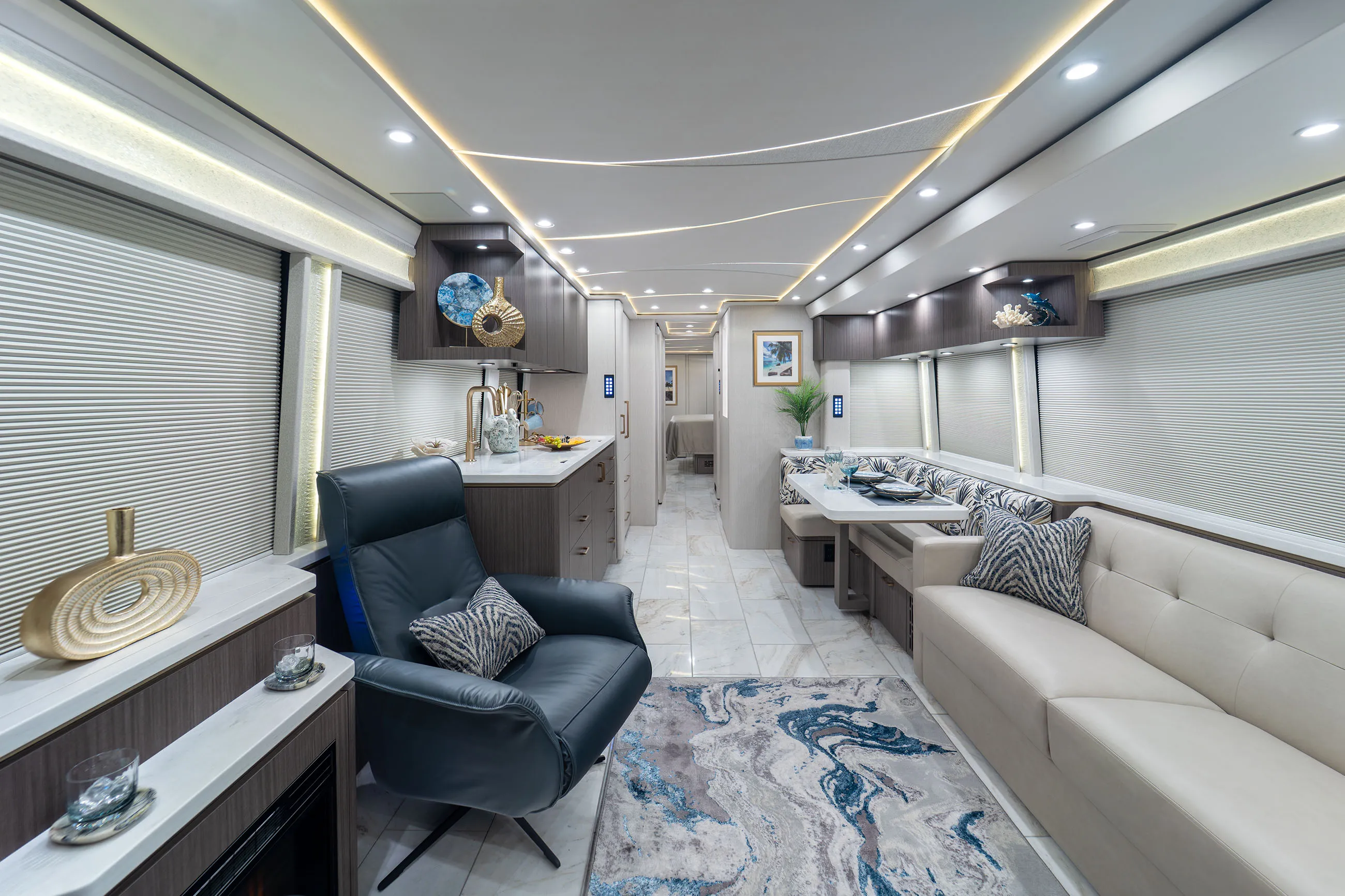 Modern RV interior with a cream sofa, dark leather recliner, dining area with patterned cushions, marble floor, and ceiling lighting.