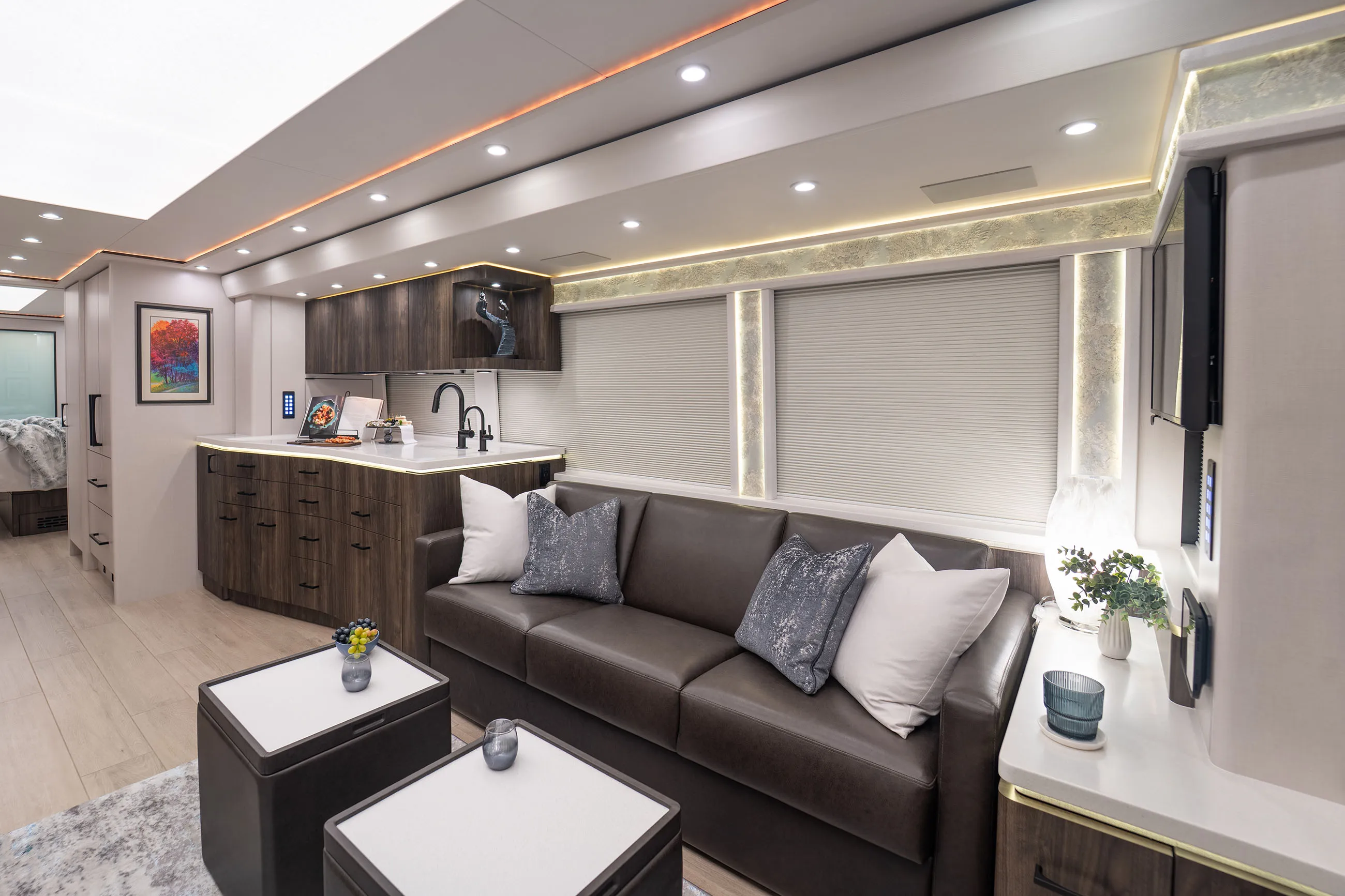 Modern RV interior featuring a dark leather sofa with cushions, a compact kitchen with dark wood cabinets, and two small white-topped tables.