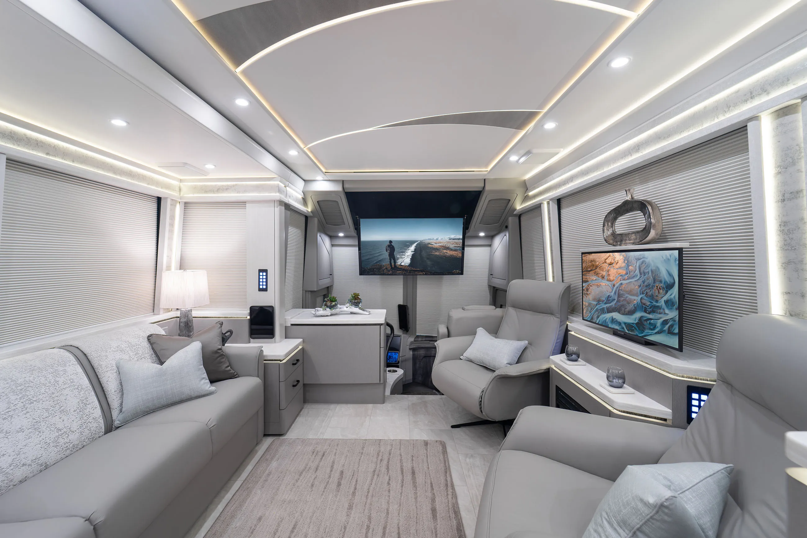 Modern RV interior with two gray leather recliners, a gray sofa with pillows, two wall-mounted TVs, and soft ambient lighting.