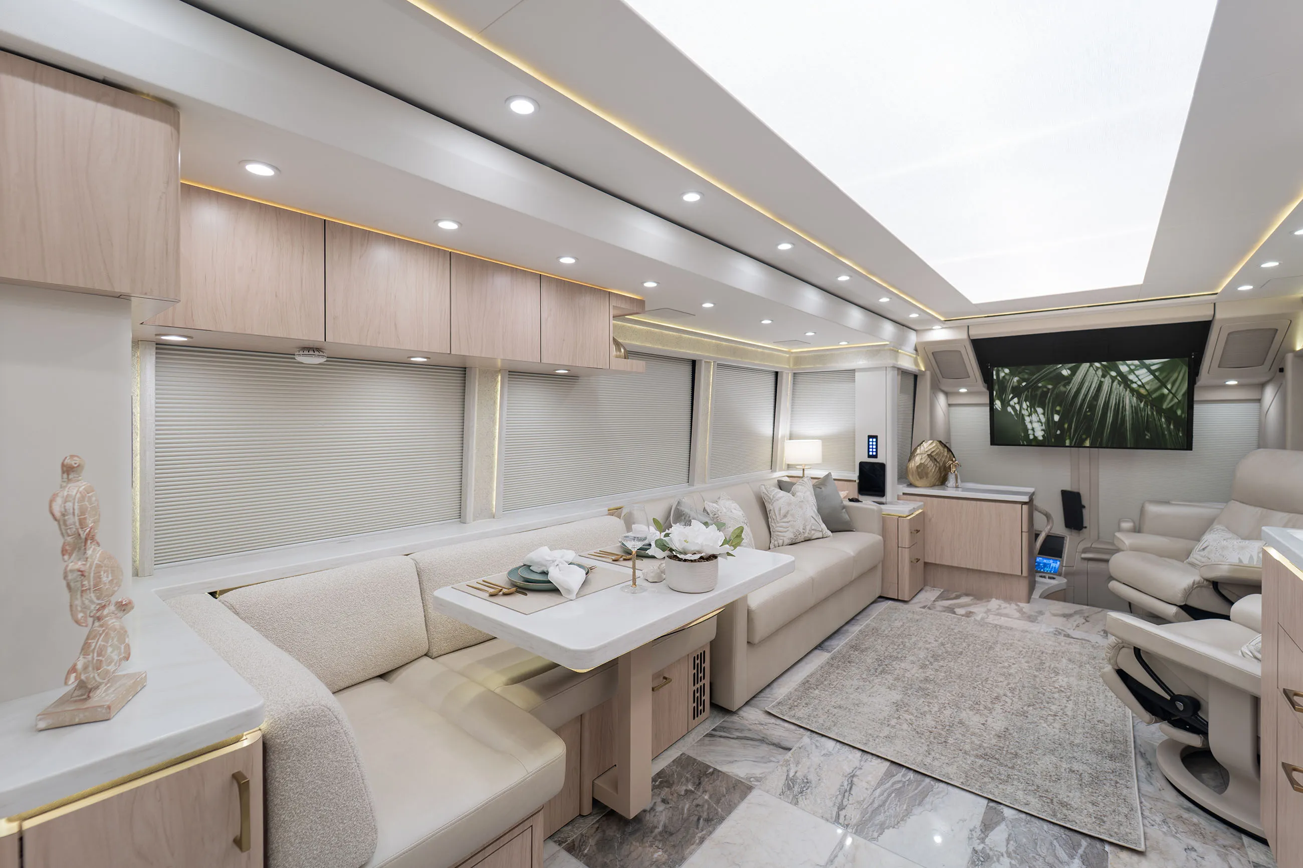 Interior of a modern RV with beige leather seating, a dining table set with plates and napkins, overhead cabinets, a large TV displaying palm leaves, and marble-style flooring.