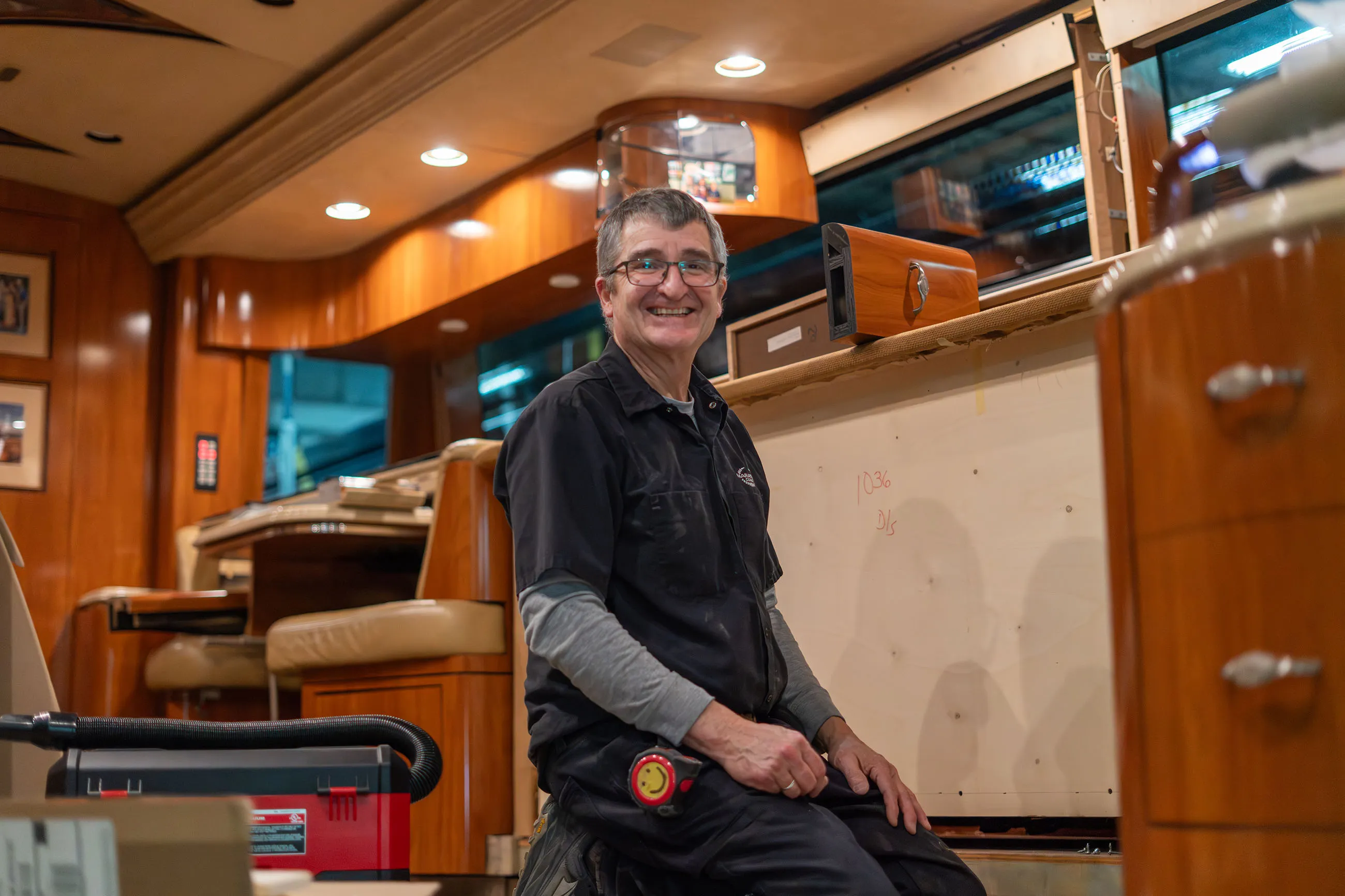 Smiling man wearing glasses and work clothes kneeling inside a wooden-paneled room with tools and equipment around.